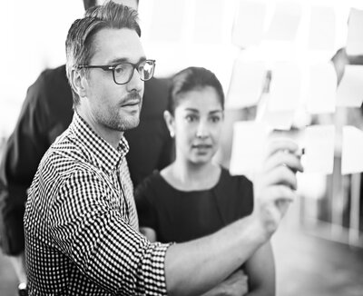 rsz_photo_man_pointing_to_sticky_notes_on_glass_wall_showing_colleagues_bw rsz_photo_man_pointing_to_sticky_notes_on_glass_wall_showing_colleagues_bw